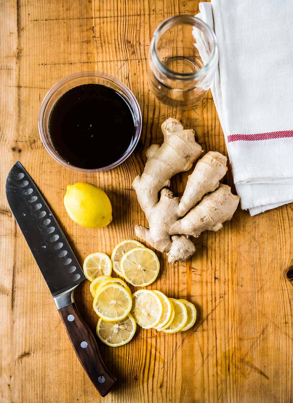 Ingredients for lemon ginger tea with honey, including raw honey, ginger root, lemon slices, whole lemon, a knife, and storage jar.