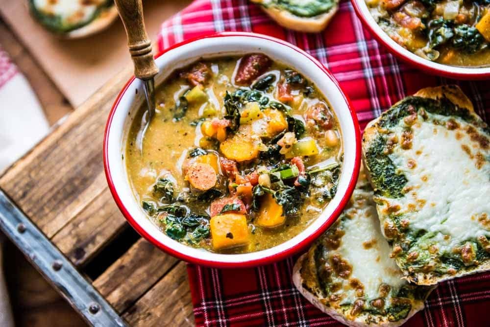 one bowl of minestrone soup sitting on a wooden crate with a picnic blanket and some pesto ciabatta bread.