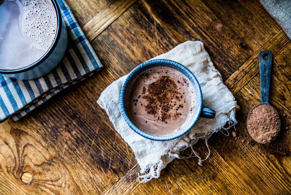 A blue mug filled with rich homemade hot chocolate sits on the countertop, accompanied by a tablespoon of cacao powder and vegan milk.