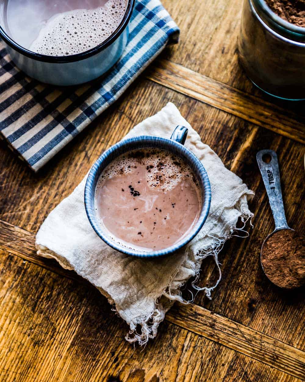 A mug of dairy-free hot chocolate rests on a cheesecloth, next to a tablespoon of cacao powder.