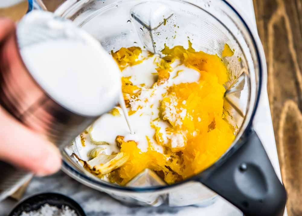 all the soup recipe ingredients in a vitamix blender container, with coconut milk being poured in.