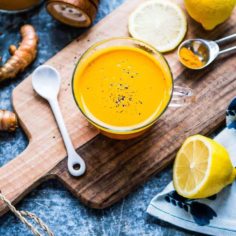 yellow turmeric tea in glass mug on a wooden cutting board surrounded by lemon and spoons.