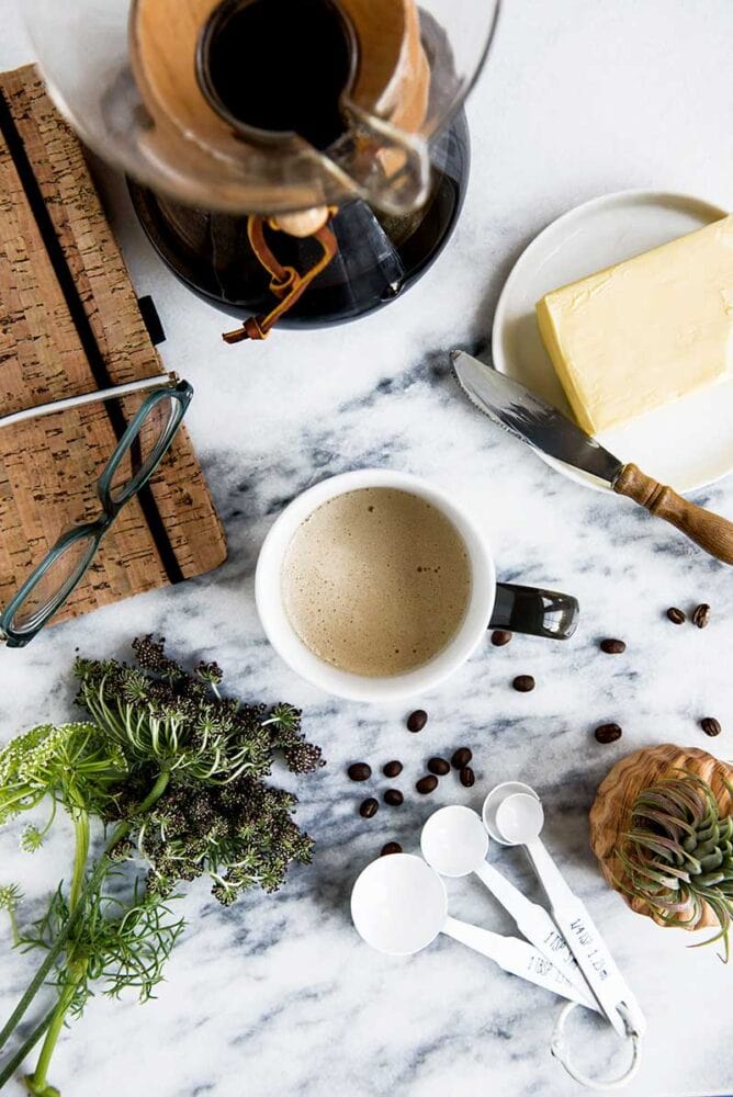 overhead shot of a cup of healthy coffee, with a block of butter and a used butter knife, loose whole coffee beans, and greenery