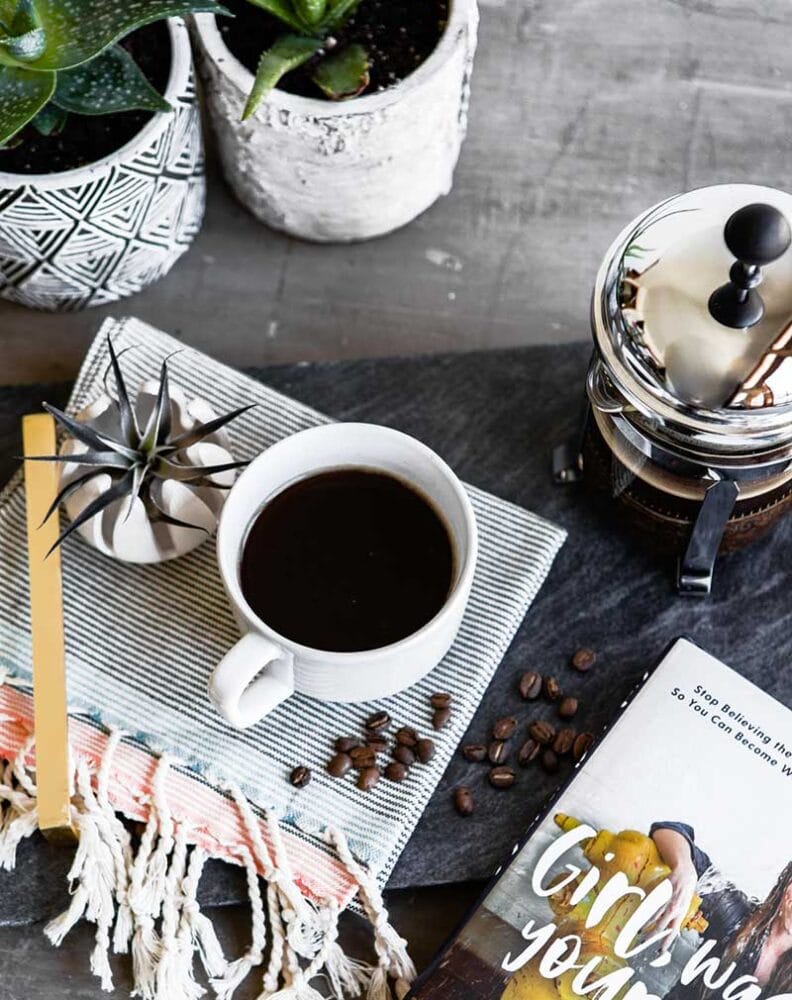 overhead shot of poured cup of healthy coffee, with french press, various succulents, and the corner of a book also seen