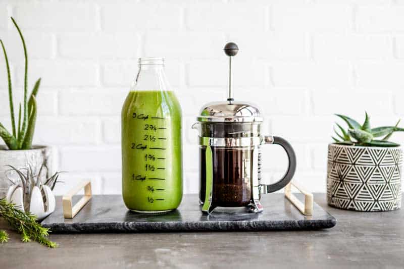 glass jug full of green smoothie next to a french press full of healthy coffee on a marble slab