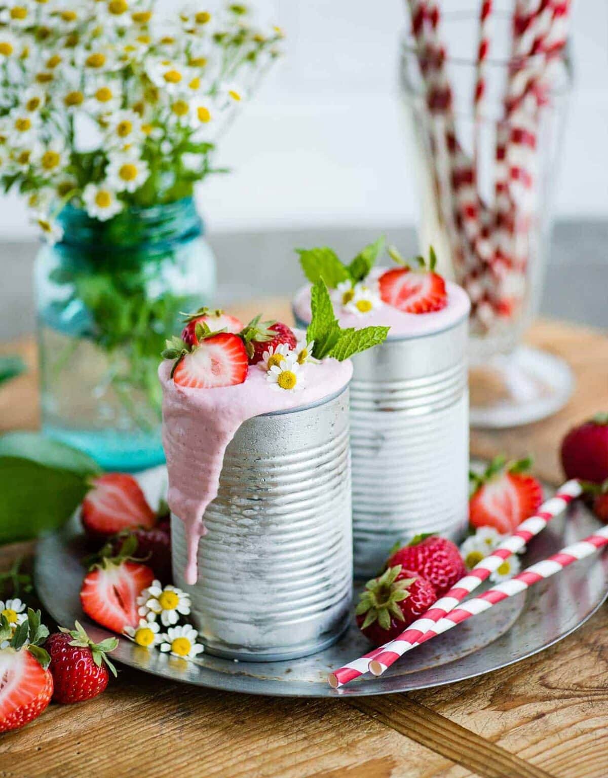 A refreshing and colorful display of two vegan strawberry milkshakes, garnished with fresh fruit and mint, with whole strawberries and straws arranged nearby.
