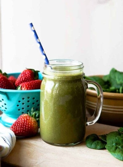 spinach strawberry banana smoothie in a glass mug surrounded by fresh spinach and strawberries.