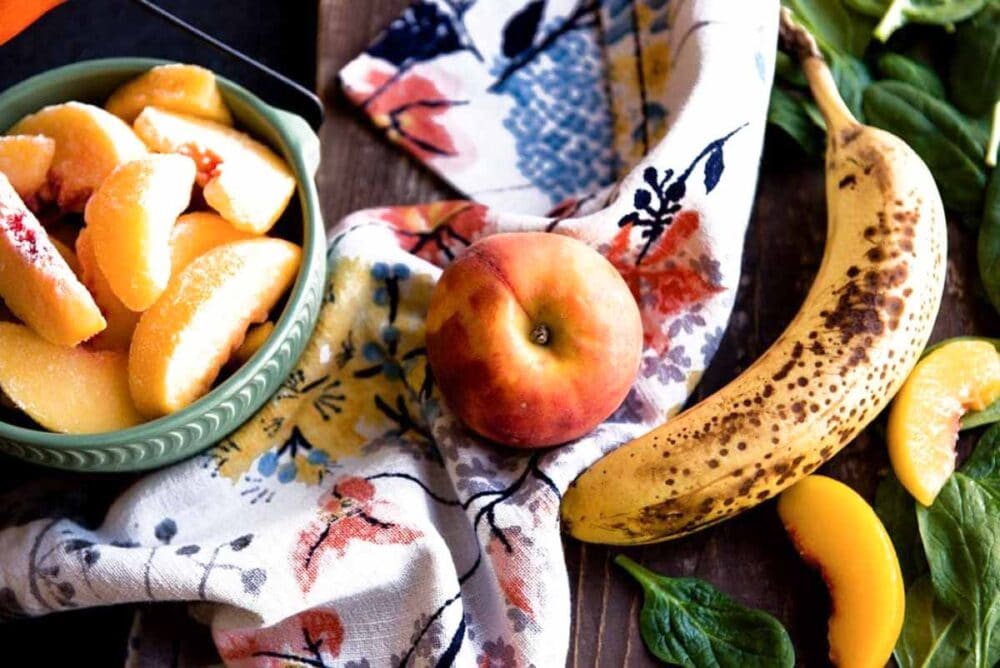 Ingredients for a vegan green smoothie on a wooden counter including peaches, spinach and banana.