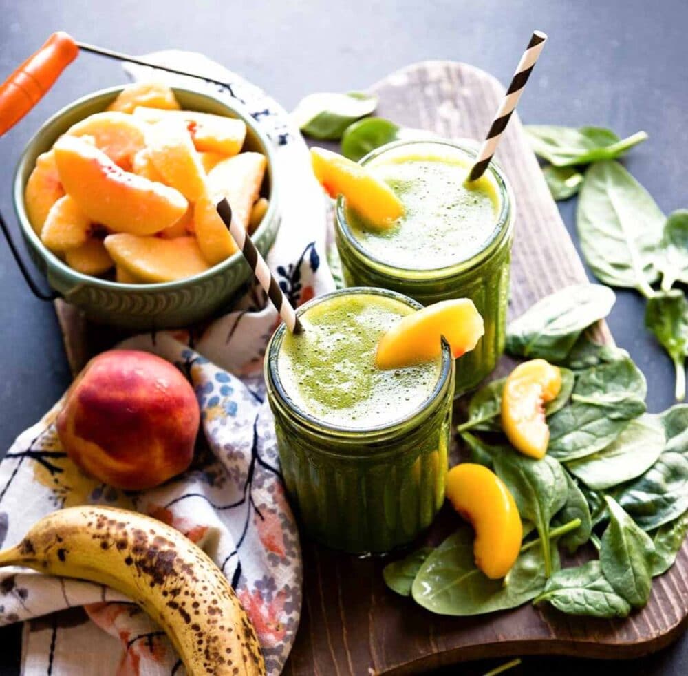 2 green smoothies in glass jars with paper straws, topped with peach slices.