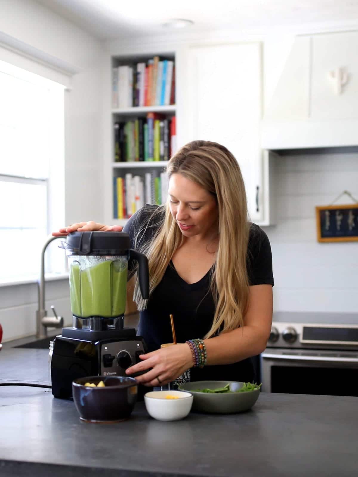 white woman using a Vitamix blender to blend a green smoothie in a kitchen.