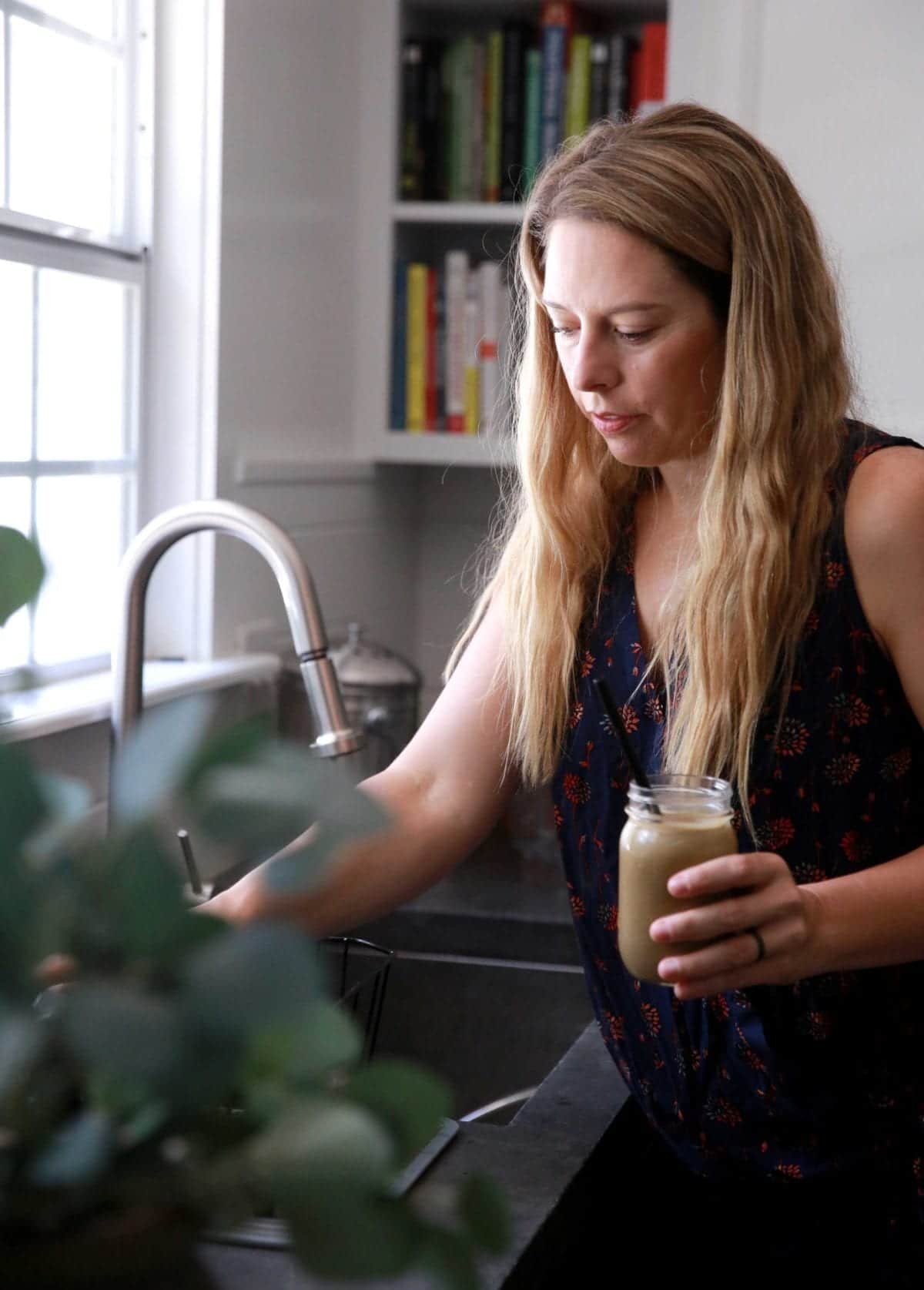 Jen Hansard standing over a kitchen sink, holding a chocolate weight loss protein shake in a glass jar with a black straw.