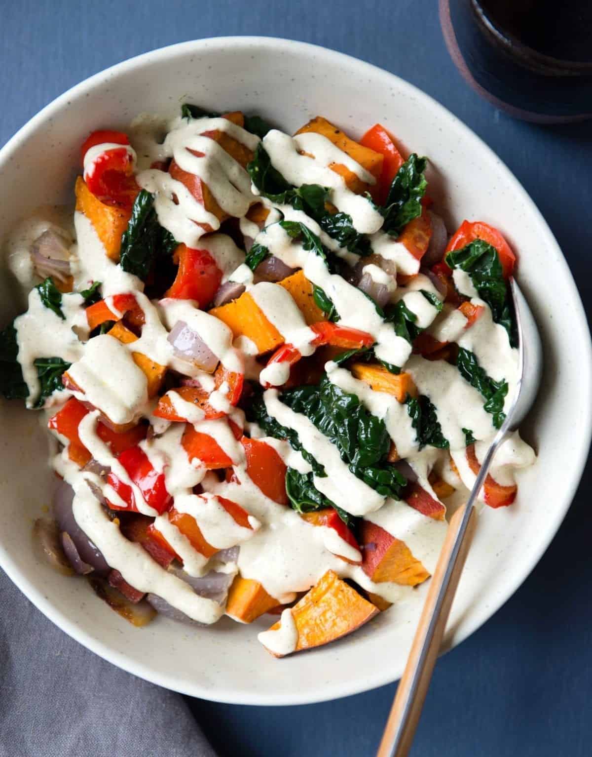 Zoomed in view of colorfull cooked veggies in white bowl topped with white hemp sauce.