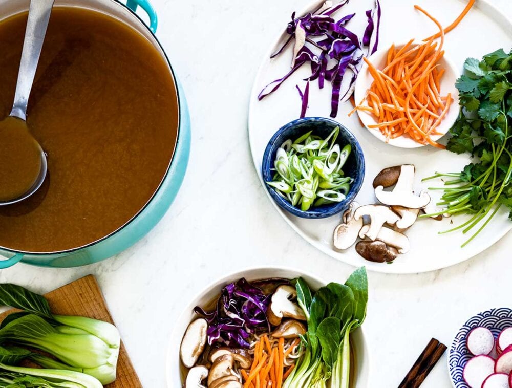 blue stock pot of vegetable stock next to a bowl of vegetarian ramen.