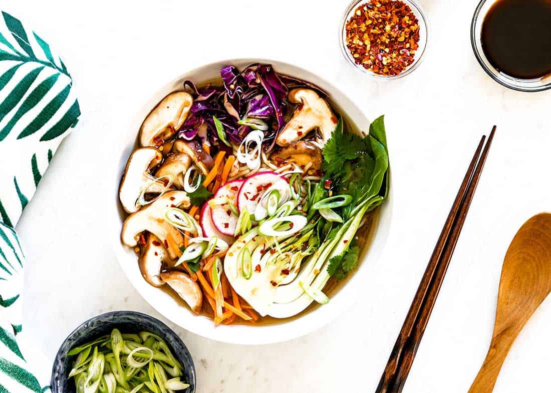 Vegetarian Ramen soup in a bowl next to chop sticks, red pepper flakes and green onions.