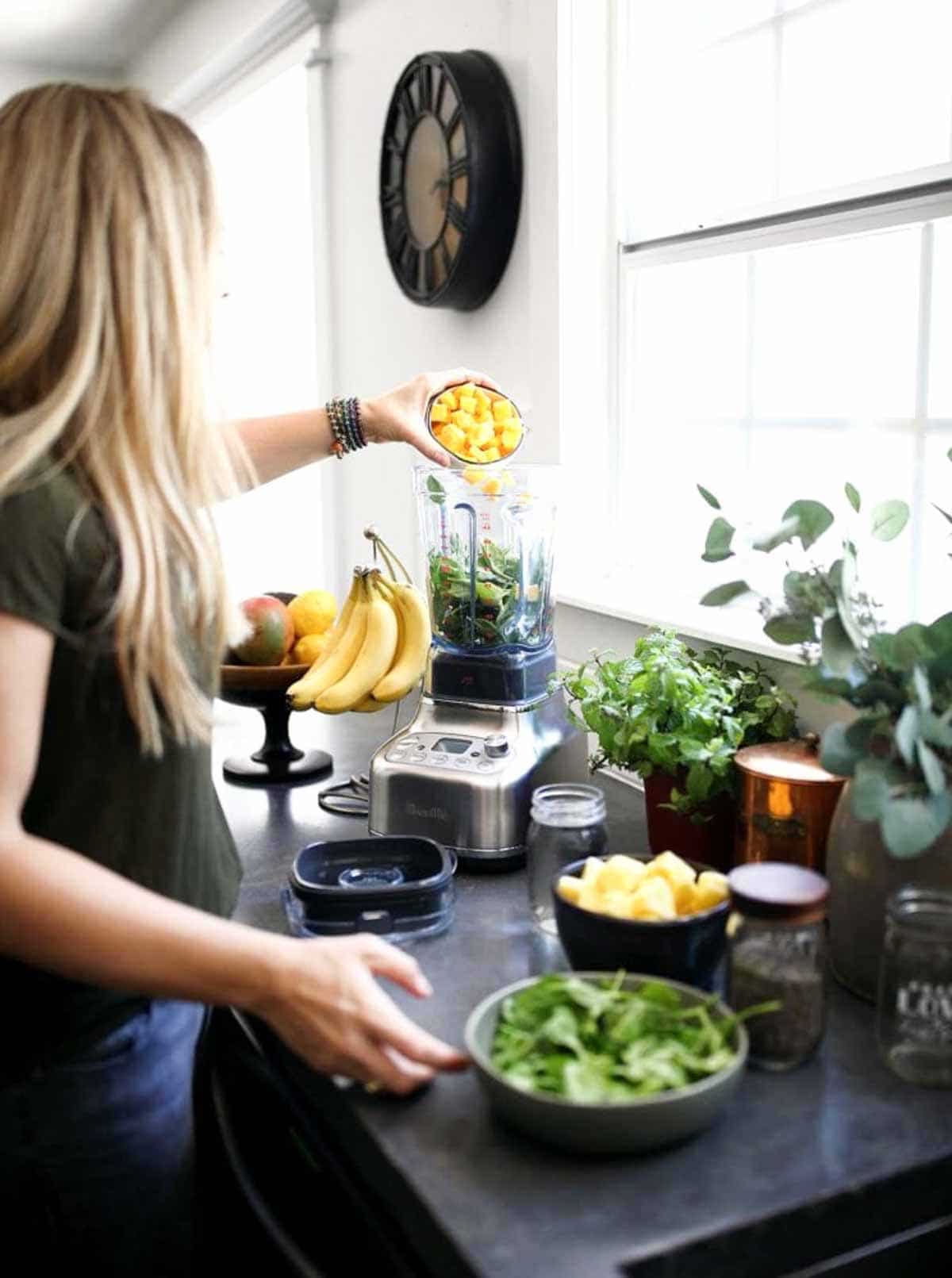 white woman with blond hair pouring chunks of pineapple into a blender to make a breakfast smoothie.