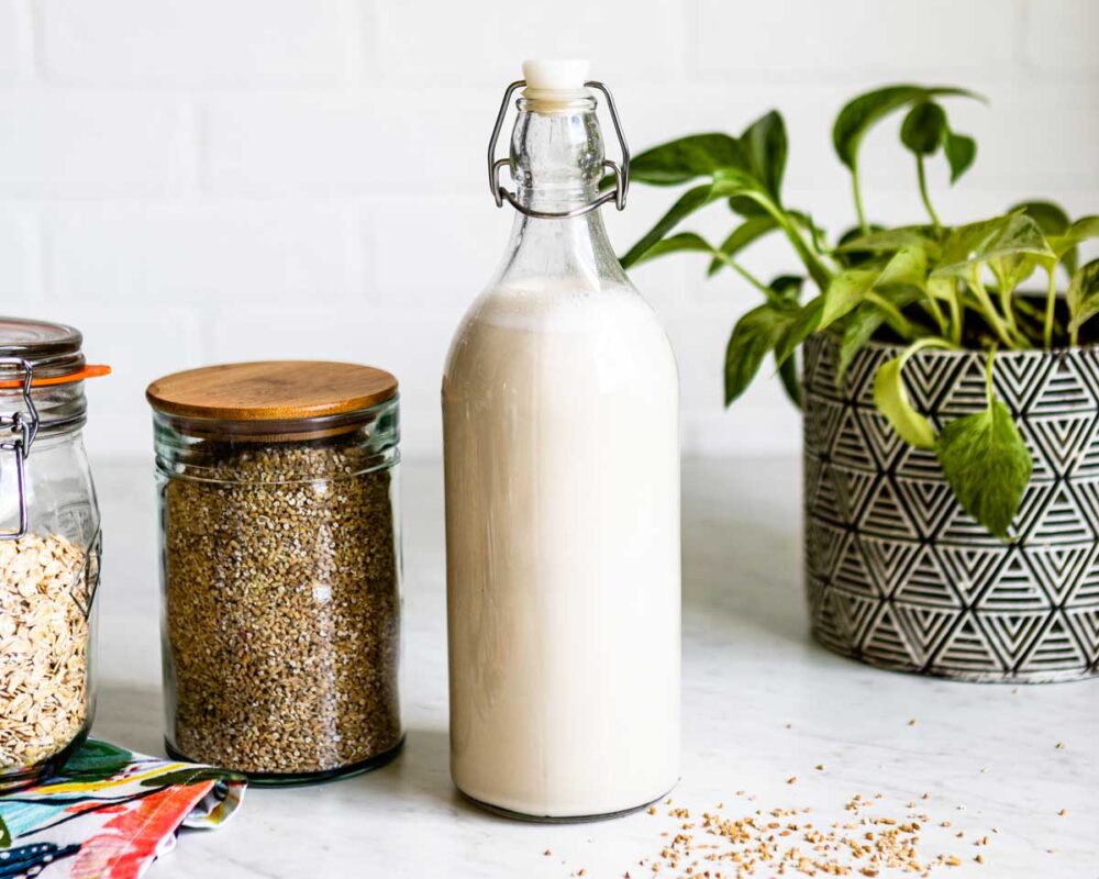 glass pitcher of oat milk next to containers of steel-cut oats and rolled oats.