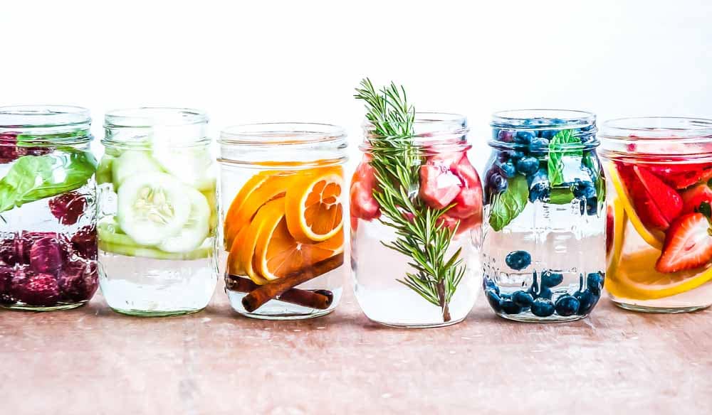 Mason jars filled with detox water in a row with berries, oranges, rosemary and grapefruiit