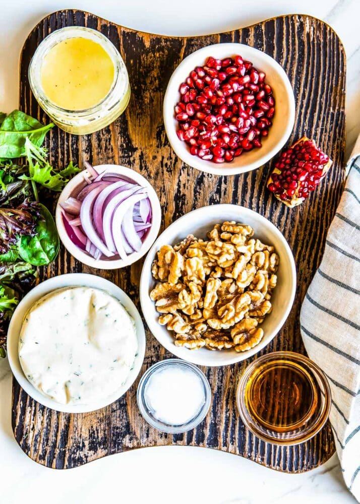 ingredients for a pear salad including vinaigrette, pomegranate arils, red onions, walnuts, vegan cheese, mixed greens coconut oil and olive oil.