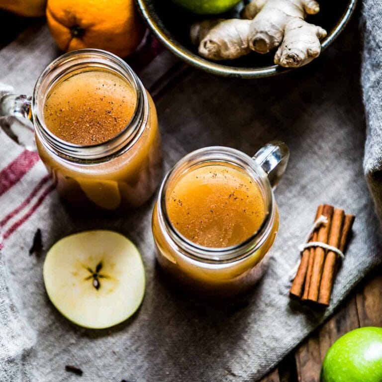 2 glass mugs of crockpot apple cider on a tea towel surrounded by fresh ingredients.