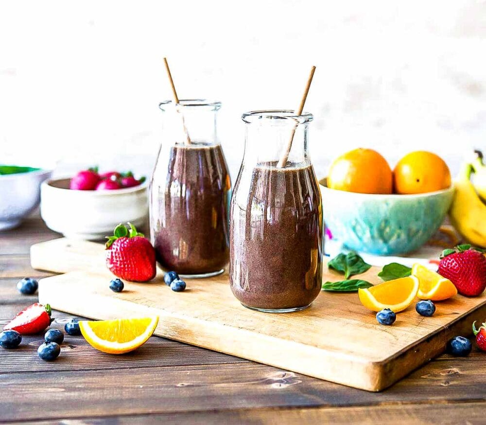 2 glasses of purple smoothie with paper straws on a table surrounded by fresh produce.