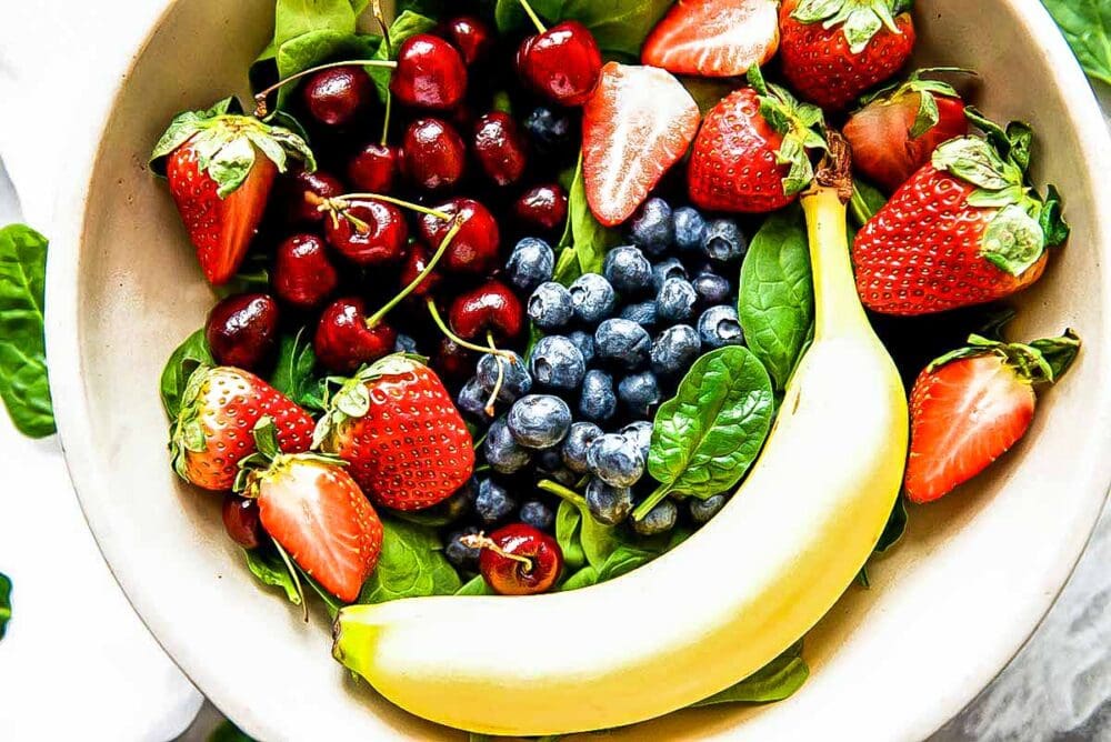 bowl of fresh ingredients including strawberries, blueberries, cherries, banana and spinach.
