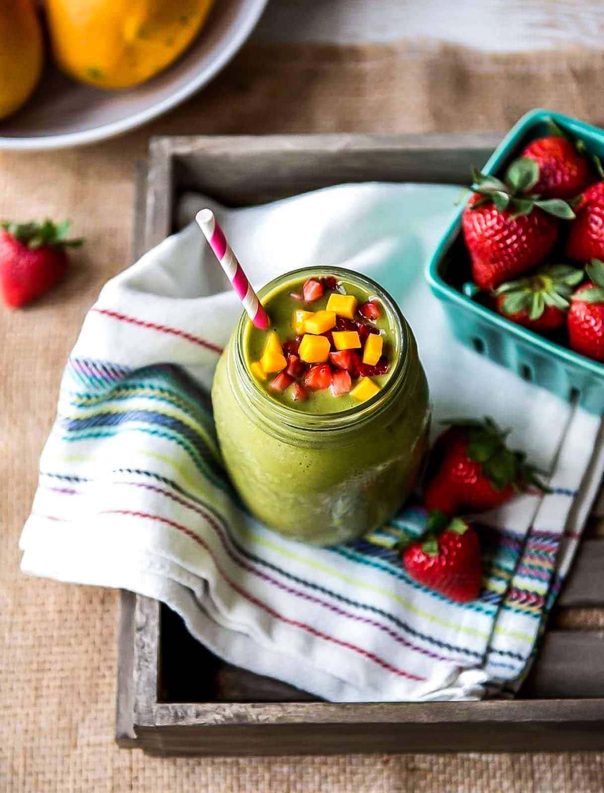 green smoothie in glass jar with paper straw topped with fresh mango and strawberry cubes.