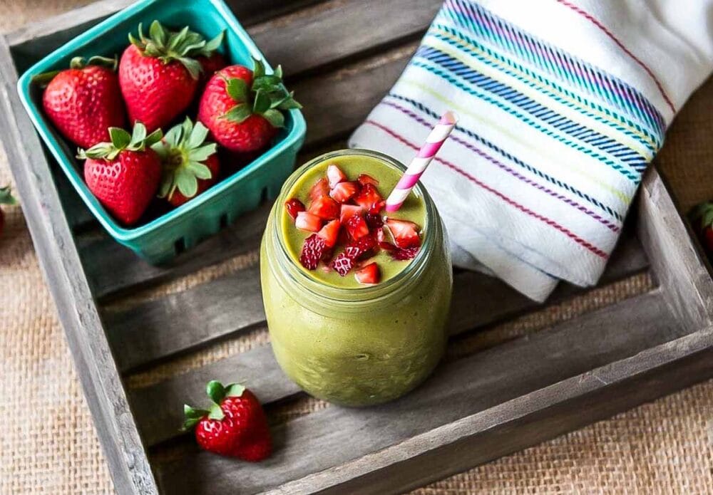 green smoothie in glass jar with striped paper straw sitting in a wooden crate next to a container of fresh strawberries.
