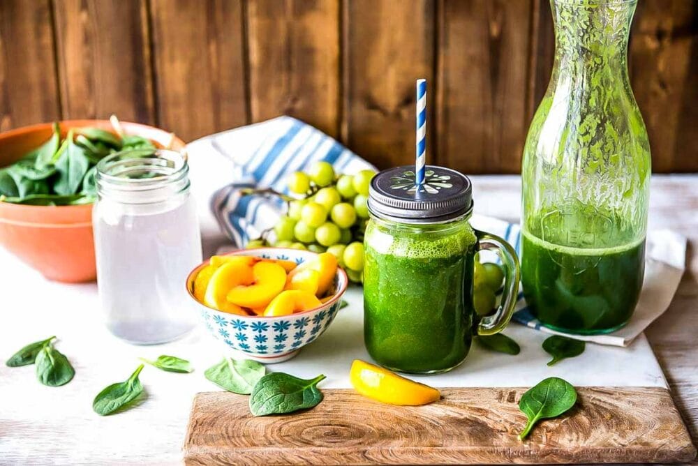 green smoothie in a glass mug next to a liter of green smoothie and bowls of grapes, peaches, coconut water and spinach.