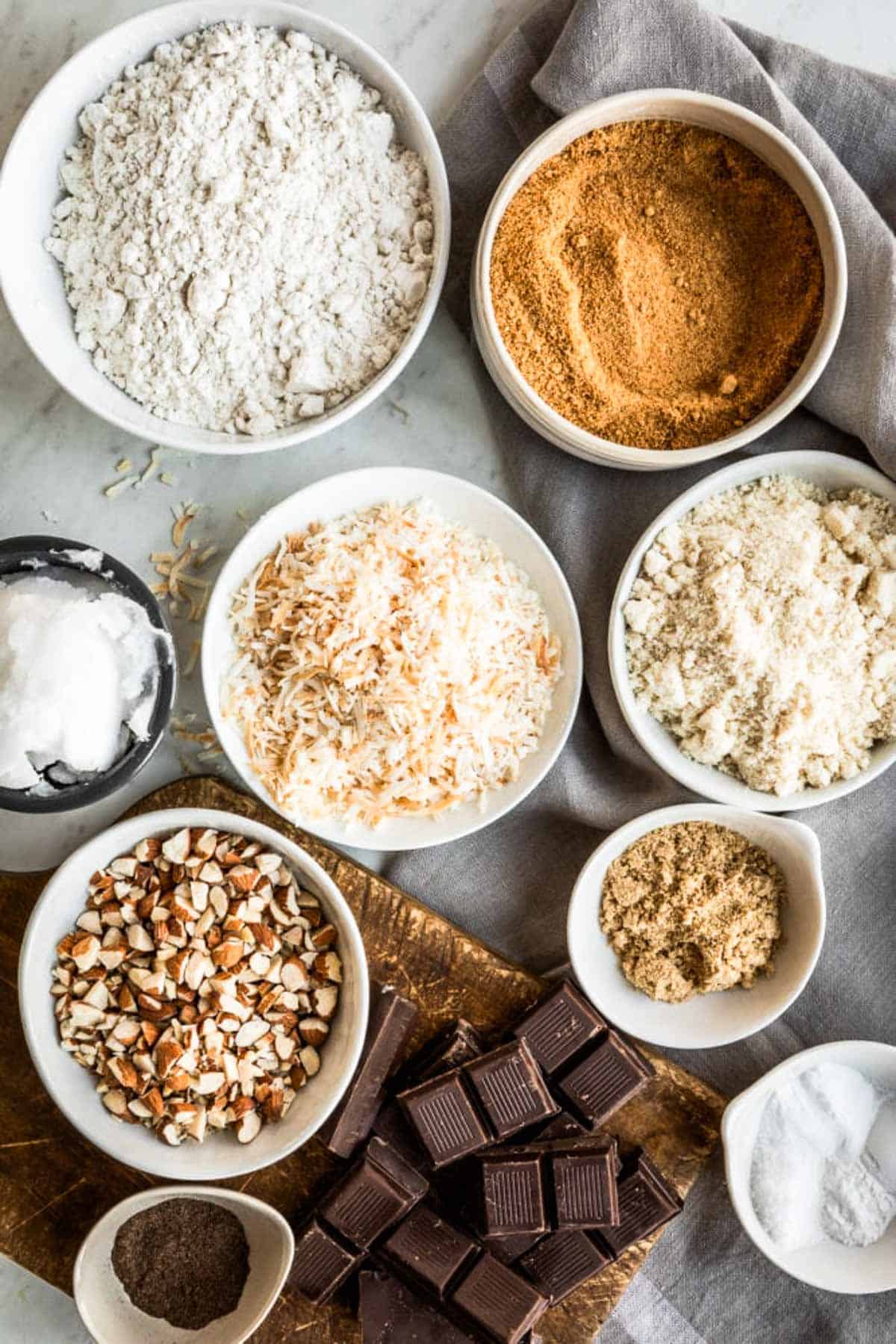 Ingredients for cookies including gluten-free flour, coconut sugar, almond flour, coconut flakes, coconut oil, chopped almonds, flaxseed, chocolate squares and sea salt.