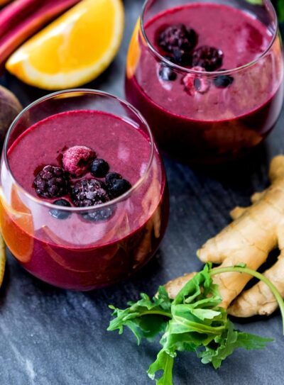 Two glasses full of one of the best smoothie recipes for anti-inflammatory, topped with berries on a countertop, surrounded by ginger root, kale lemon slices, and beets.