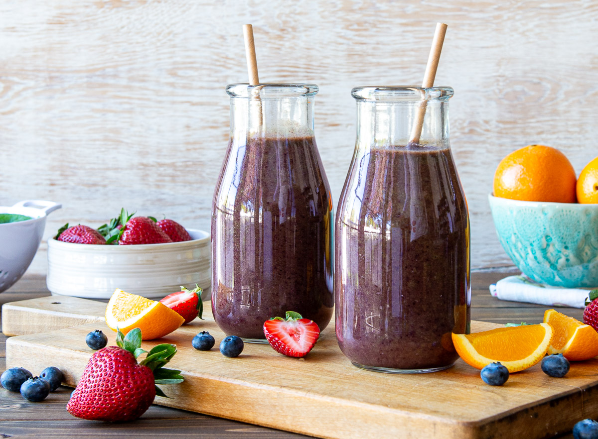 2 glasses of purple smoothie with paper straws on a table surrounded by fresh produce.