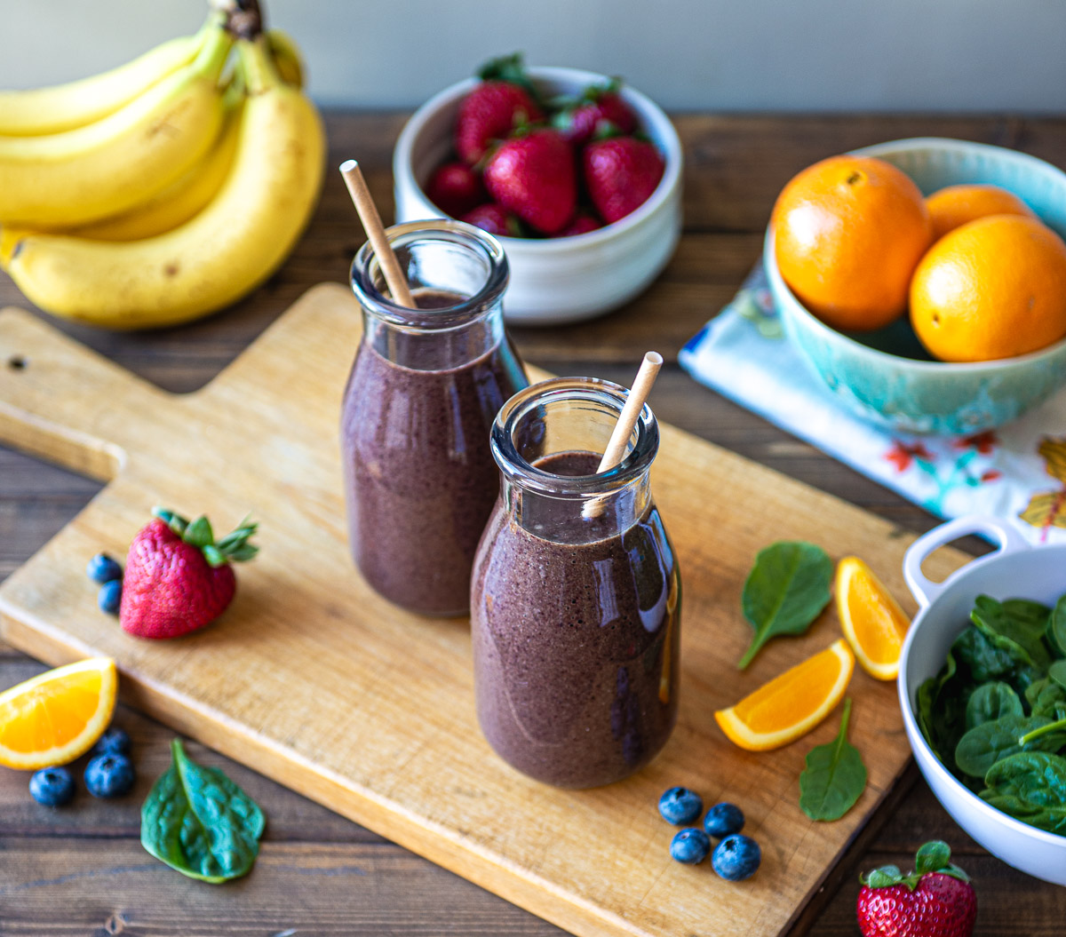 Two glasses of purple smoothie with paper straws on a table, surrounded by fresh produce.