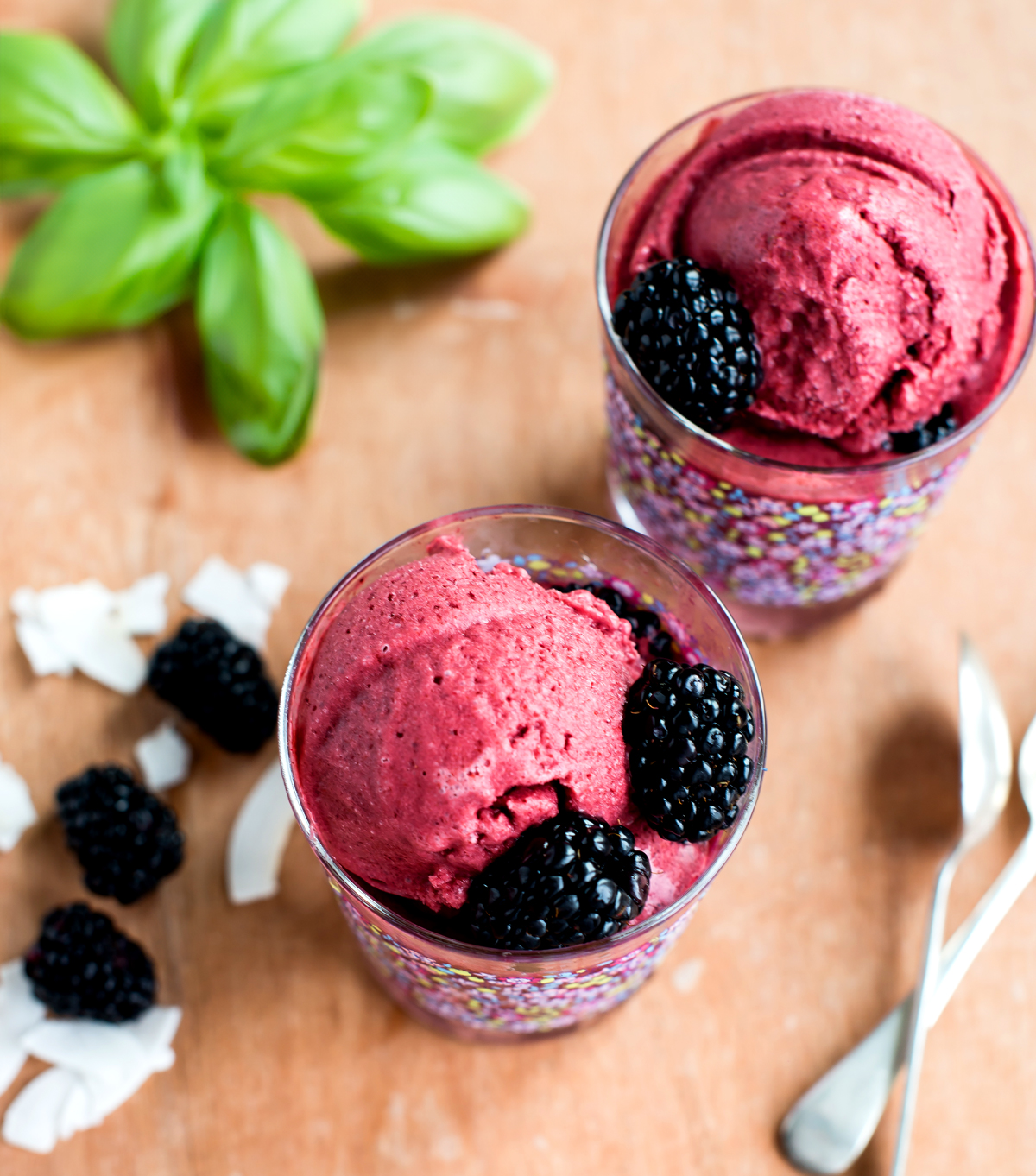 Two glasses filled with refreshing homemade blackberry coconut sorbet, displayed with blackberries, coconut flakes, and silverware on a woodgrain countertop.