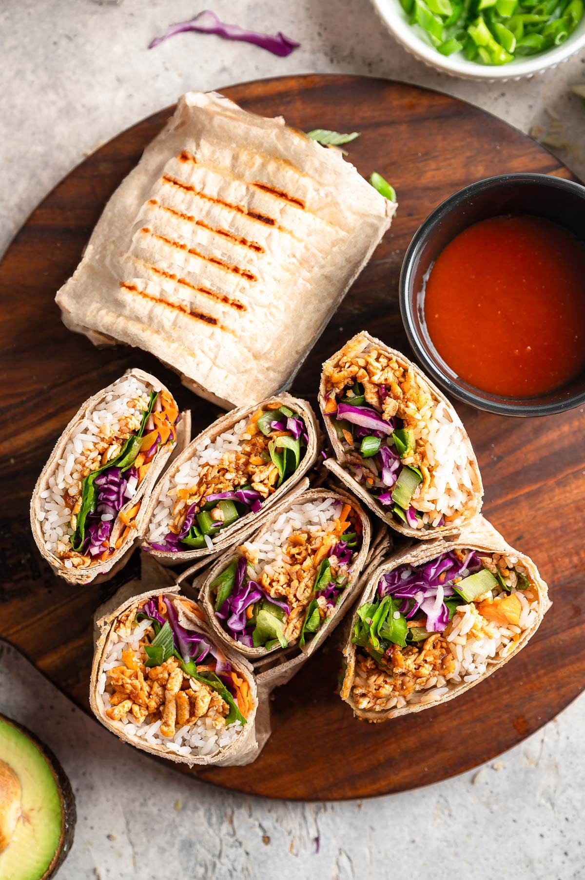 A wooden board displays a vibrant arrangement of buffalo shredded tofu wraps with buffalo sauce on the side.