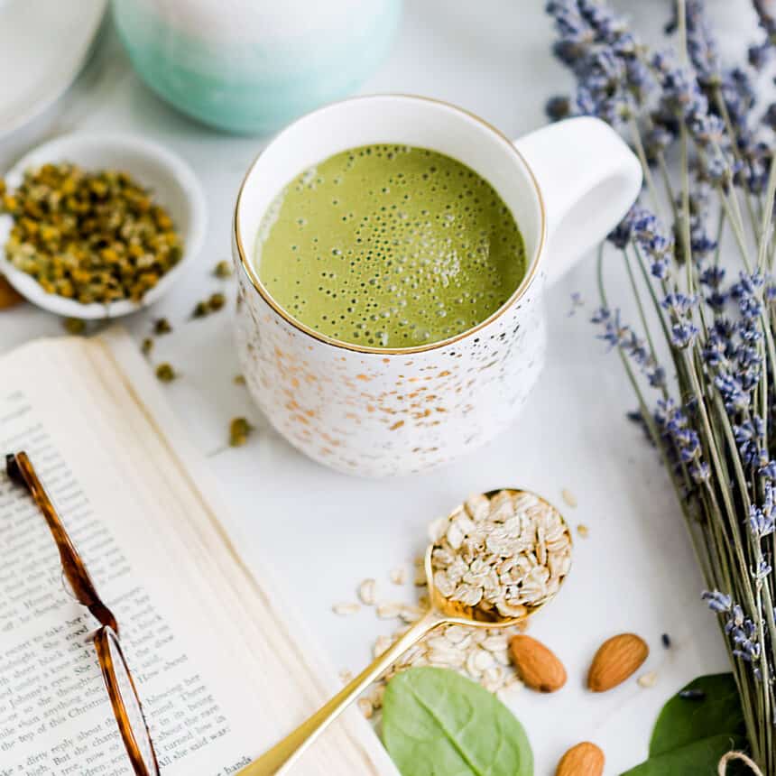 Green bedtime smoothie in a white mug surrounded by lavender, almonds, tea, and spinach.