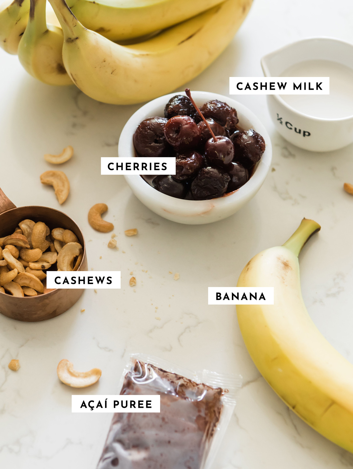 Ingredients for an acai smoothie recipe, including bananas, frozen cherries, cashew milk, cashews, and a packet of acai puree, arranged on a marble countertop.