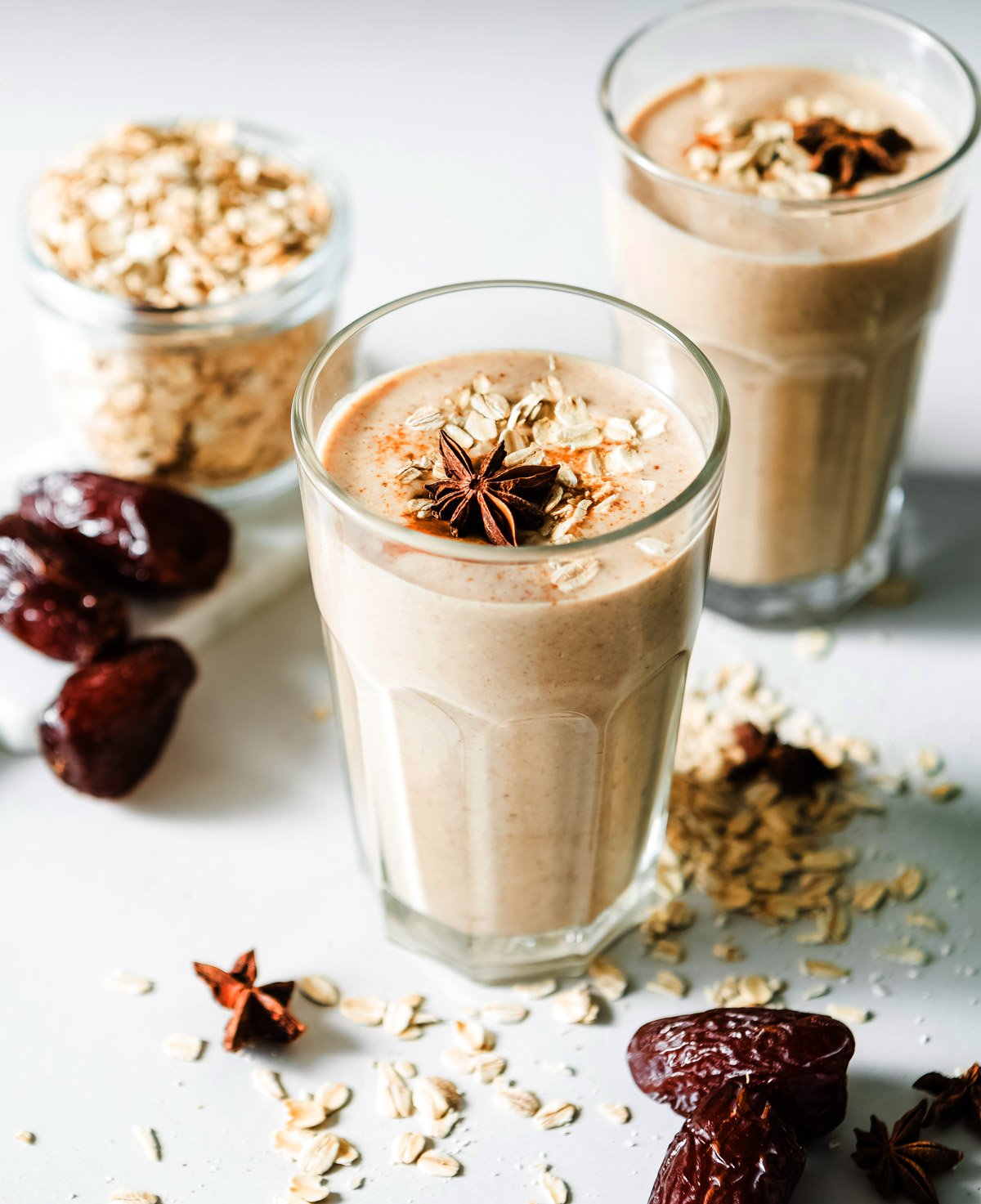 Glass of date smoothie topped with rolled oats and star anise, with oats and dates scattered on the countertop.
