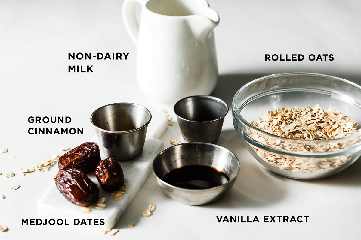 Bowls of Medjool dates, rolled oats, vanilla extract, and milk, arranged on a white countertop to make a smoothie.