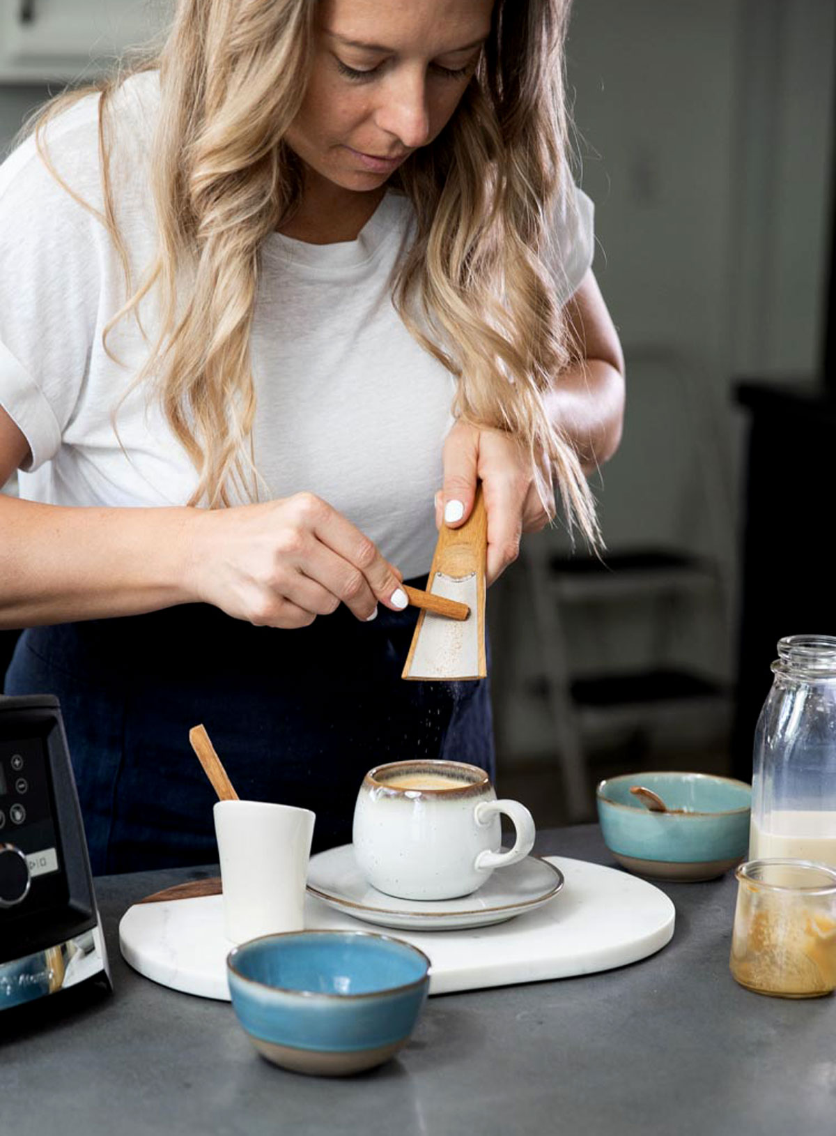 Woman grating fresh cinnamon into a mug of cinnamon dolce latte, surrounded by latte ingredients.  