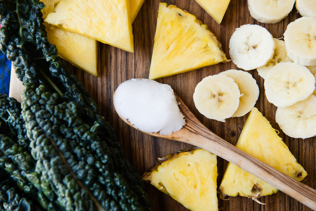 a wooden spoon filled with coconut oil, surrounded by fresh ingredients like kale, pineapple, and banana.