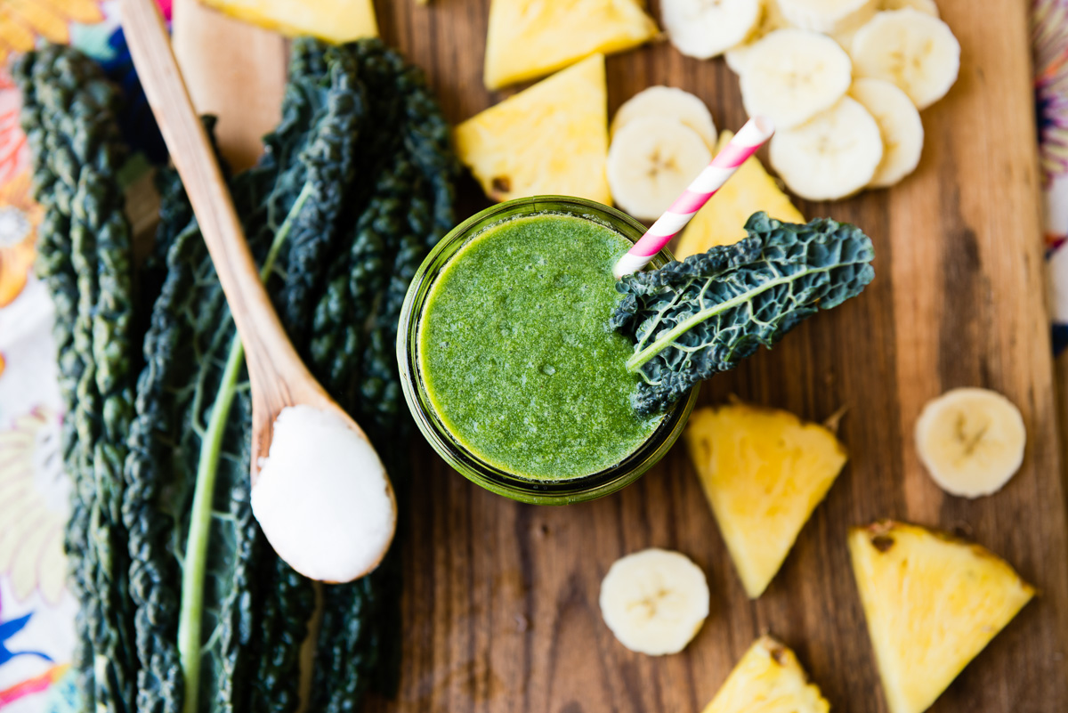 Top-down view of a coconut oil smoothie garnished with a kale leaf, surrounded by fresh ingredients.