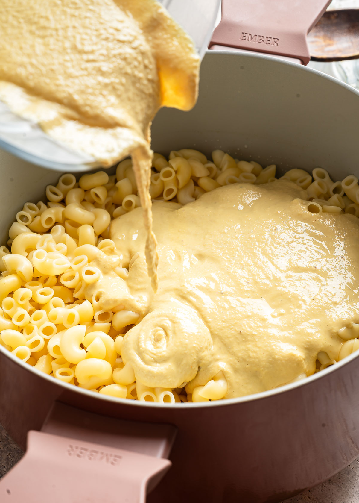 Creamy white vegan cheese sauce being poured over cooked elbow pasta