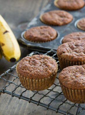 Several gluten-free banana muffins on a cooling rack and in a muffin pan.