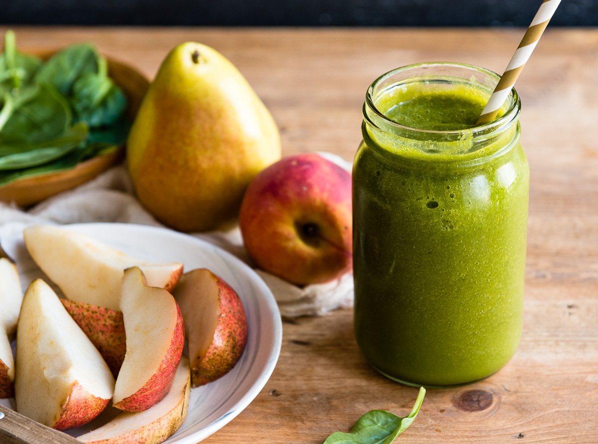 Pear smoothie in a glass, surrounded by sliced pear, peach, and fresh spinach leaves