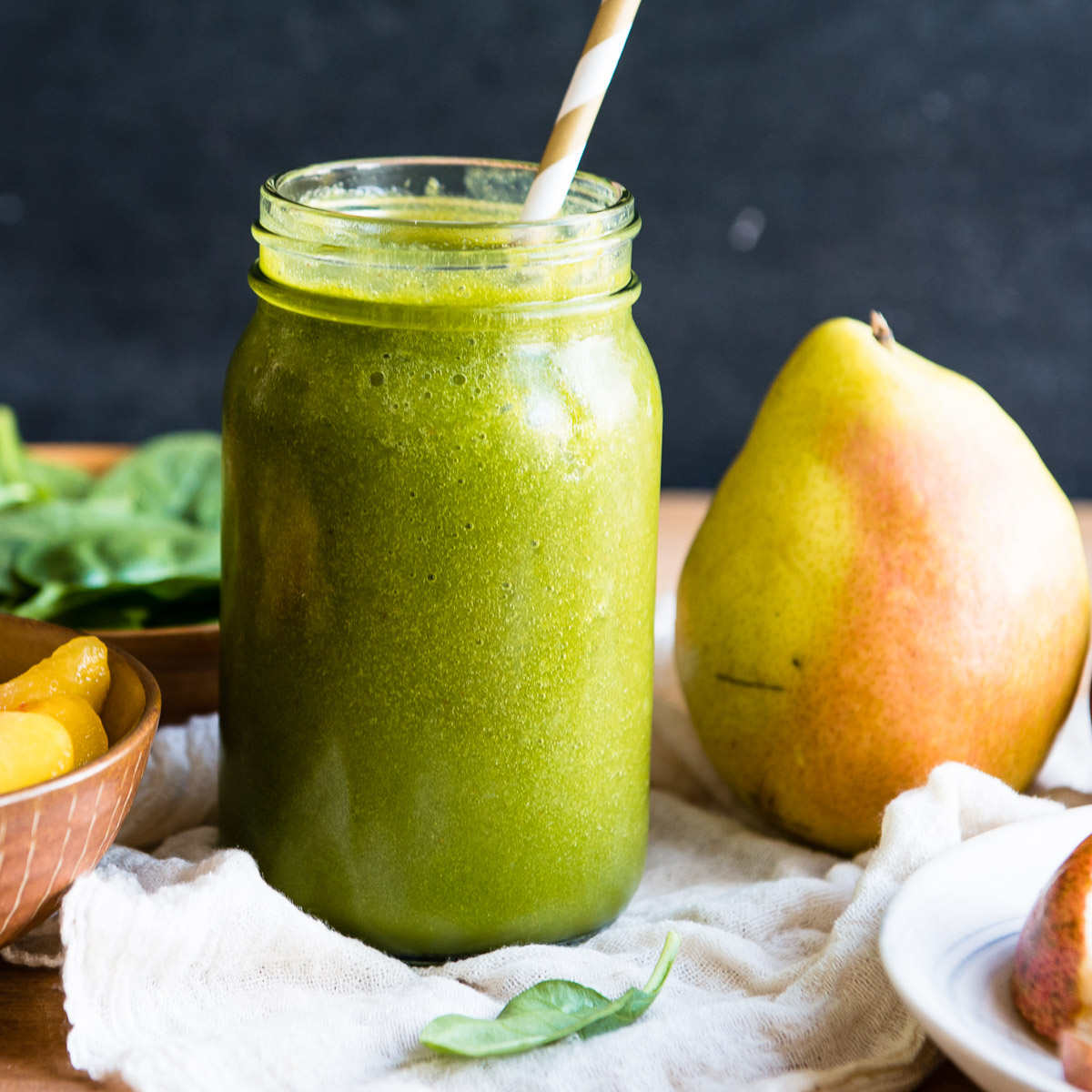 large glass jar with green smoothie and a paper straw, next to peach slices.
