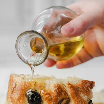 A small glass container of olive oil being poured onto fresh, baked bread.