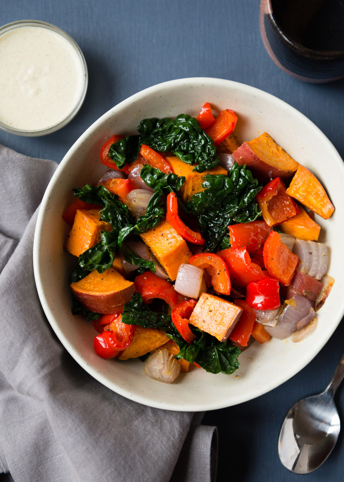 White bowl filled with roasted sweet potatoes, bell peppers, red onions, and kale, served with a spoon on a gray napkin.