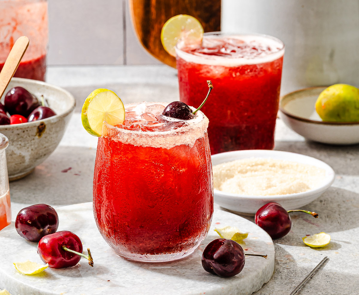 A vibrant red cherry limeade in a stemless wine glass, with a garnish of a cherry, a lime wedge, and a sparkling sugar rim.