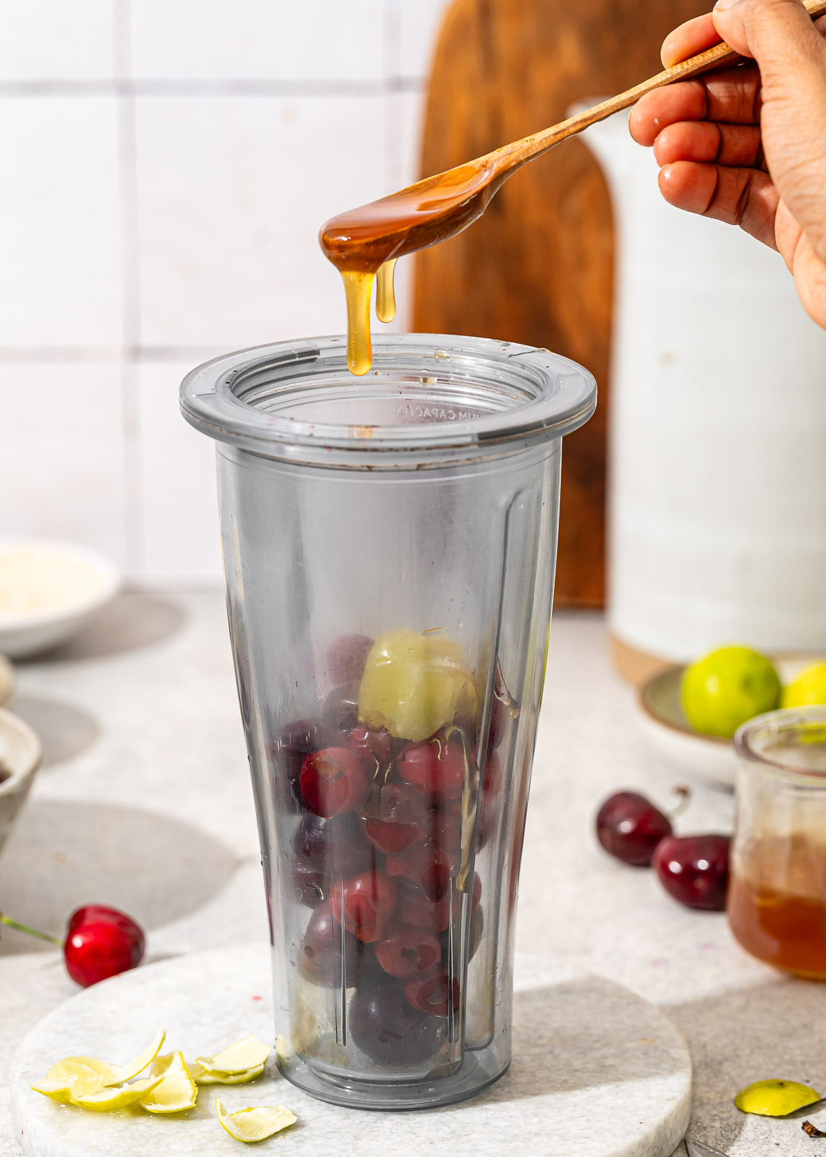 A close-up shot of honey being drizzled from a spoon into a blender filled with cherries and limes.