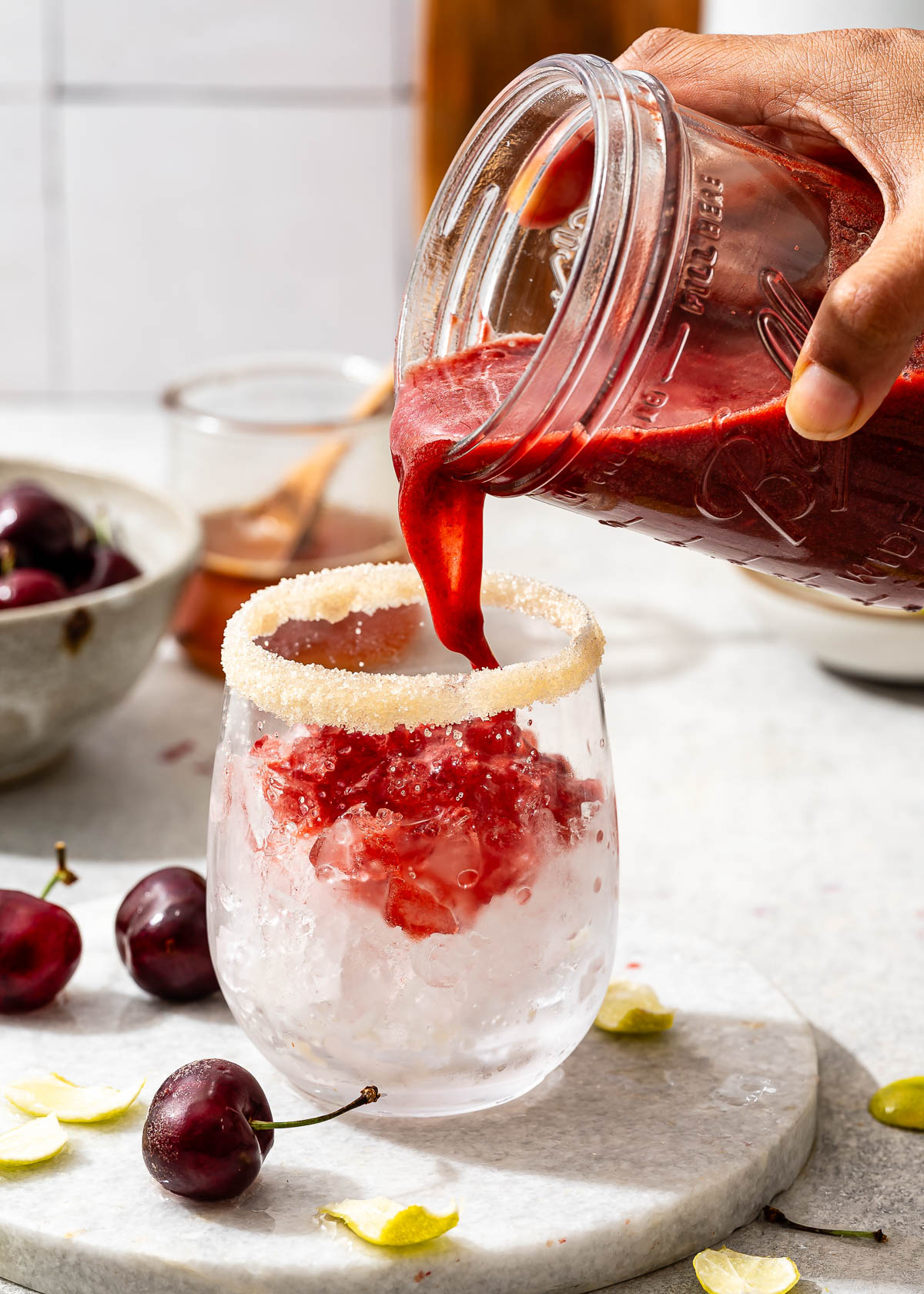 A hand pouring a cherry limeade mixture from a jar into a stemless wine glass with ice and a sugar rim.